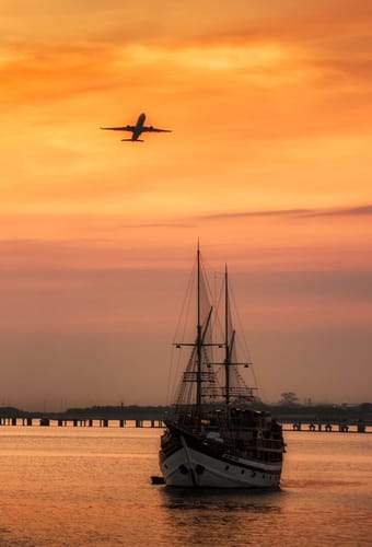Solnedgang over Benoa havn på Bali med sejlskib i forgrunden og fly, der letter i horisonten